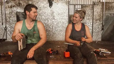 Aaron Magee Aaron Magee with short brown hair is sitting on a stage to the left. He is wearing a dirty green vest top which is covered in sheep dirt and sweat. He is sitting beside a girl with blonde hair in a black vest top, she also has dirt on her. She is holding shearing equipment. There is also equipment beside them. There are silver metal panels behind them.