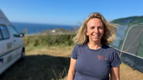 A woman stands in a field overlooking the coast, she is in between a tent and a campervan.