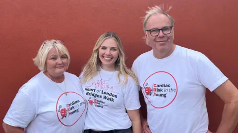 BBC Two women and a man are stood together in front of a red wall. They are all smiling and wearing white T-shirts that display a Cardiac Risk in the Young logo. The first woman, on the left of the picture, has short blonde hair and a pair of glasses on top of her head, the second woman, in the centre of the picture has long blonde hair and a T-shirt that also displays the words: Heart of London Bridges Walk. The man on the right of the picture has short grey hair and is wearing glasses.