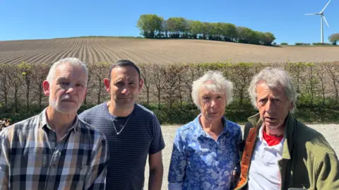 Lee Trewhela / LDRS Three men and a woman stand in front of a field. A trimmed hedge separates the road from the field. Green trees are in the distance. A large wind turbine is located behind the field. The sky is blue.