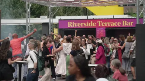 BBC A crowd of people dance and socialise outdoors in front of the Riverside Terrace Bar at London’s Southbank Centre. The atmosphere appears lively, with many smiling, raising their arms and interacting under a metal-framed canopy.