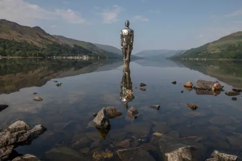 Alec Davies Mirrored statue of a man standing in a still loch with some large rocks poking out towards the shore and surrounded by grassy hills