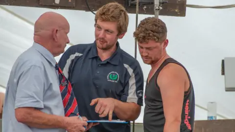 Lucymoosh Aaron Magee is standing in the middle wearing a navy t-shirt. He is pointing at a clip board which has a sheet of paper on it. He is looking at a man with a bald head wearing a light blue shirt and red, white and blue tie. There is also a man in a black vest top looking at the clip board. The background is white. 
