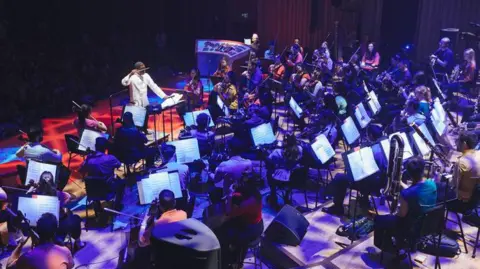 Giulia Spadafora A conductor dressed in white leads a large orchestra. The picture is taken from behind the musicians, so you can see their open sheet music, and they are lit by dark blue and purple lighting. 