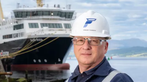 Ferguson Marine Graeme Thomson in a white hard hat, wearing glasses, with a large ship in the background