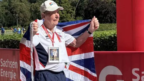 Karen Piotr An 89-year-old man wearing Team GB sports uniform and a white cap, holding a union flag behind him, and wearing a photograph of himself and his wife around his neck.