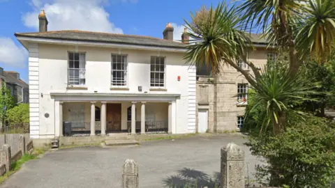 Google Maps The Redruth Albany RFC clubhouse is a white two storey Georgian style granite house with three long windows on the upper floor. There are two columns each side of the wooden entrance door and a chimney on each side of the roof. A tarmac car park is empty in front of it which is outlined with carved granite pillars. There are palm trees in the hedge lining the car park. Attached to the club house is a three storey granite building.