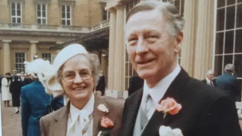 Harkness family Jack and his wife Betty stand outside Buckingham Palace smiling at the camera, with other people dotted around behind them. He wears a black suit jacket with a salmon coloured rose in the lapel, and a white shirt with silver tie.