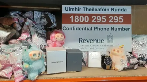 Revenue Irish Tax and Customs Blue, yellow and pink Labubu dolls next to a sign for Irish Revenue. Watches and their boxes are also present.