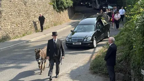 A.P Mcdonald Funeral Services A black hearse being led by a man wearing a tuxedo suit and top hat, leading a brown donkey along the road.