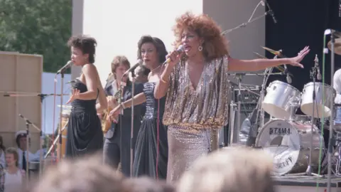 Mark Hodgson A mid-shot of three women performing on an open-air stage in bright sunshine. One woman to the right of frame is slightly closer to the camera, wearing a glamorous, silver-gold sparkling dress, and singing into a handheld microphone held in her right hand. She is about 40 and has slightly ginger-tinged dark hair in a large bouffant. To the left of frame and slightly behind on stage are two other singers in black dresses and with black hair, both slightly younger-looking, singing into microphones on stands.