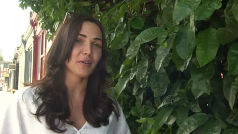 Vasilisa Ermakova, a woman with long dark hair and wearing a white top, standing next to the tree