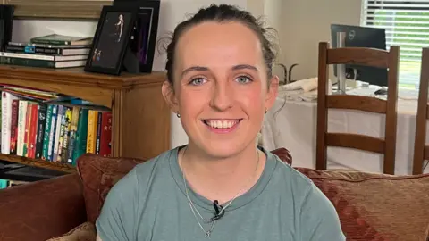 BBC A young woman, Amy Symington, smiles broadly at the camera. She is sitting in the living room of a home. In the background, through an entrance into another room we can see a dining table. There is a also a low bookcase filled with books and topped with family photos. Amy has dark hair tied back and is wearing a light green t-shirt.