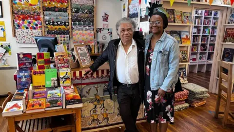 BBC A man and a woman are stood in the middle, in front of a cashier desk in a bookshop. The man is on the left and has grey hair. He is wearing a white shirt, unbuttoned at the collar, and a black jacket, with black trousers and a belt. He is leaning against the desk with his right arm. The woman on the right is wearing a black hair bonnet, and is wearing a black dress with red and white floral decorations and a light blue denim jacket. To their left, there is a table displaying a colourful selection of books. There is a computer and more books on the desk, and behind the desk there is a display of wrapping paper hung on the white wall. There are more book shelves behind the two people, on their right.