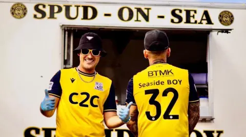 Danny Last Bradley Gibbs and Max Linduschka are standing in front of their Spud-on-Sea van which is white and has the name at the top. Mr Gibbs is on the left looking towards the camera, and Mr Linduschka is on the right with his back towards the camera. They are both wearing a yellow and black T-shirt.