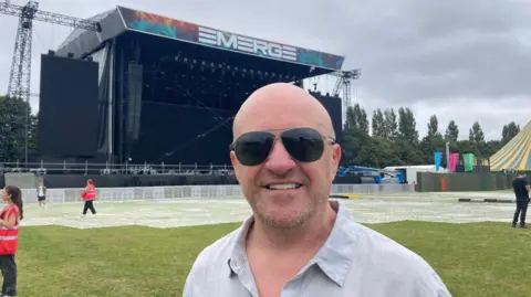 Alan Simms wearing sunglasses and smiling at the camera, he is wearing a grey shirt. Behind him is a large stage with a sign reading 'EMERGE' above it. There are a number of trees to the right of the stage, and a tree to the left of the stage. The sky is overcast. There are a number of people in the background, some of whom are wearing high-vis jackets. 