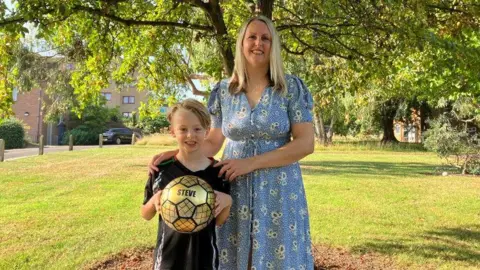 BBC A blonde, white, seven year old boy smiles in a black football kit and holding a golden football with "steve" written on it in front of a tree, next to his mother who has long blonde hair and is wearing a blue floral dress. 