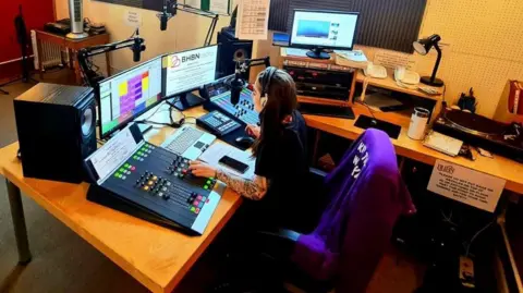 BHBN A women sits on a large chair at a desk with radio broadcasting equipment in front of her. She is looking at two screens and touching buttons on the mixing desk. There are microphones, a record player, speakers and, lights and other equipment around the desk.