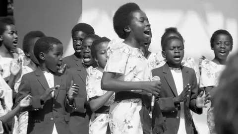 Mark Hodgson A black-and-white mid-shot of a group of approximately a dozen children, clapping and singing. One child, a girl slightly taller than the rest, is roughly central to the image, with a hand-held microphone in her right hand. The girls wear pale dresses with a floral pattern, while the boys wear dark jackets.