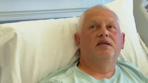BBC A man with grey hair wearing a light green t-shirt lies in a hospital bed, propped up against a pillow. He is looking upwards.