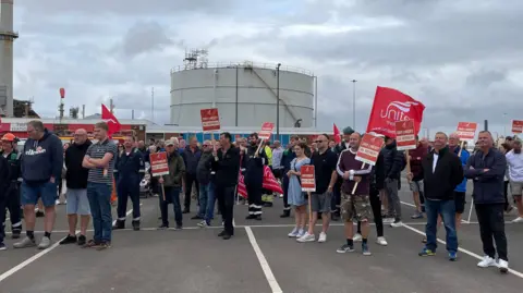 BBC A group of people gathered in the car park of Lindsey Oil Refinery. They are holding red Unite the Union signs. The sky is overcast. 