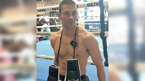 BBC A man standing in front of a boxing ring, which has a blue floor. He is topless with a pair of black and white boxing gloves hanging around his neck. The image cuts off at the waistband of his black pair of shorts.