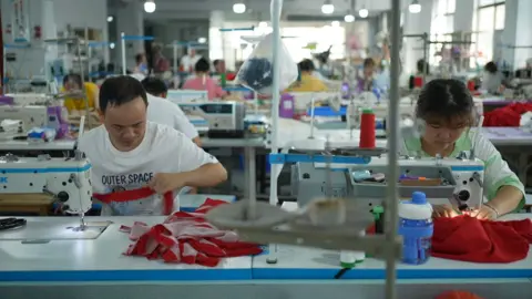 Xiqing Wang/BBC A man and a woman at a Shein factory, sitting side-by-side and working on sewing machines. They are cutting and sewing red fabric.