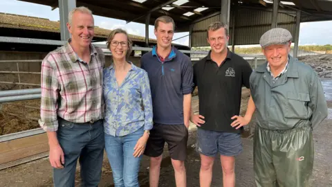 BBC Andrew Jones with wife Jayne, sons Harvey and Oscar and father Roger are standing in a line in a barn. They are all smiling at the camera.