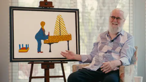 Image courtesy of Castle Fine Art A photo of Sir Billy sitting next to the artwork. Sir Billy has a full grey beard and grey hair. He has round-rimmed glasses. He wears a patchwork shirt in shades of grey. He is sat in a floral armchair in front of picture on an easel. The picture shows a stylised piano with loads of striped lines in orange and yellow. A stylised performer in a stripy blue suit has one leg on the keyboard. Behind him sits an upturned stool.