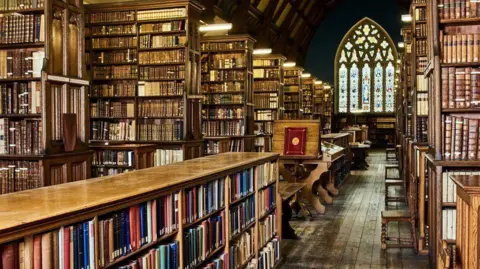 Ushaw Ancient looking wood-panelled library with rows and rows of books and stained glass window in background.
