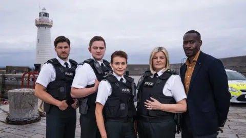 Hope Street actors Cameron Cuffe, Finnian Garbutt, Kerri Quinn, Tara Lynne O'Neill and Marcus Onilude in Port Devine. 
They are standing in from of a lighthouse and a police car.
Four are wearing police uniforms of white short-sleeved shirts, dark trousers and a dark protection vests saying Police.
Marcus Onilude, stood at the far right, is wearing a dark blazer and a yellow shirt.