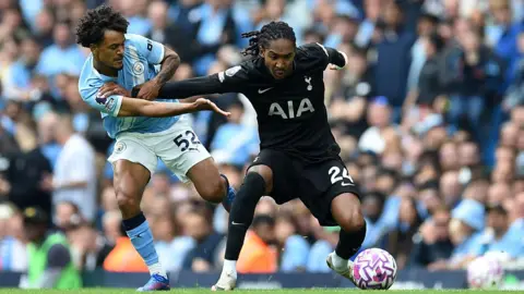 Reuters Manchester City's Oscar Bobb and Tottenham Hotspur's Djed Spence grapple with each other, with the ball at Spence's feet. Bobb is wearing the sky blue strip while Spence is in his club's black away kit. City fans watch on from a packed stand in the background.