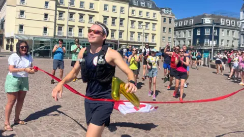 BBC James crosses the finish line of a marathon in an urban square, triumphantly holding a golden inflatable number '7'. A red ribbon marks the finish, held by another person. Behind James, other participants cheer and clap, with tall buildings and a clear blue sky in the background