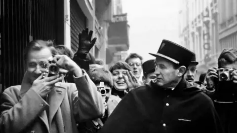 Paul McCartney, Courtesy Gagosian Titled: Photographers, fans, and officers, rue de Caumartin, Paris, January 1964. A black and white image of people looking at the camera, some are smiling, others are pointing cameras at the photographer, McCartney.