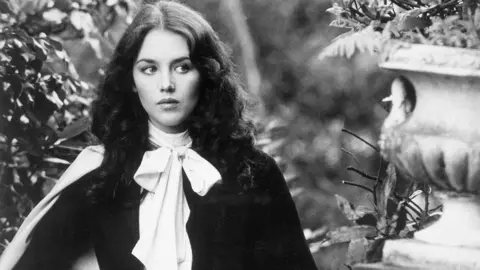 Getty Images A black and white image of Isabelle Adjani who has brunette hair and is standing in a garden with stone ornaments. 