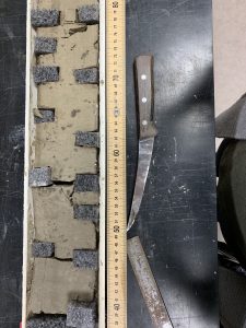An overhead photo of a black lab bench with light brown sediment, a brown yardstick, and a knife 