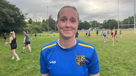 Jade Hutton with her hair tired back wearing a blue Yate RFC top. She is standing on a rugby pitch with women training behind her.