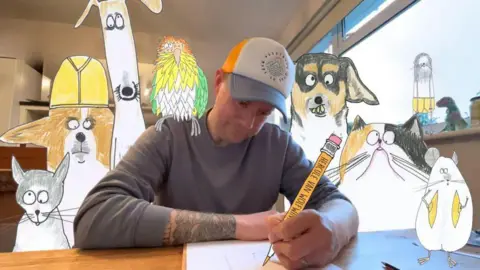 Phil Heckels Phil Heckels sat wearing a cap drawing holding a large cut out pen with large cardboard cutouts of some of his artwork surrounding him.