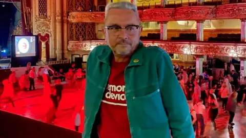 Sharon McMahon a man with grey hair and glasses wearing a green jacket and red top.