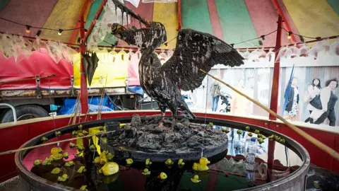 Getty Images The traditional hook-a-duck game at fairgrounds, but with  twist. Below a colourful striped canvas tent, there is a large white seabird perched on rocks in the centre of a vat of dark oil, which it is partially covered in. Its wings are outstretched and its beak is open, with crushed drink cans crammed inside its mouth. Below it are small rubber ducks which have overturned in the grease. It is likely a commentary on the damage oil spills can inflict on ecosystems and wildlife. 