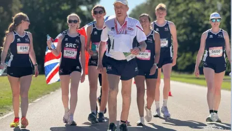 World Transplant Games An 89-year-old man who has won four medals at the World Transplant Games, he is running with six younger competitors running behind him holding a union flag.