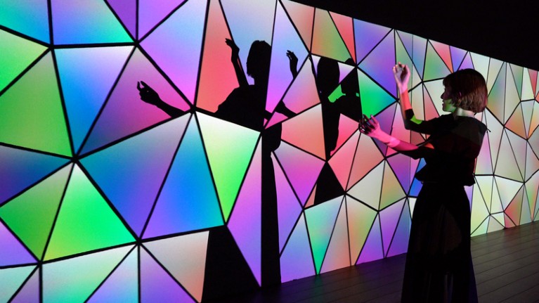 A woman holding up her arms watches how her shadow is fragmented and displayed on a wall with a brightly coloured pattern of triangles