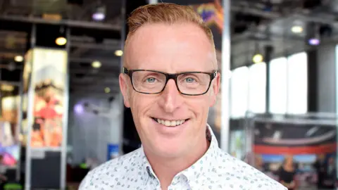 BBC Bryan Burnett smiling straight at the camera. He has short fair hair. He is wearing dark-rimmed glasses. He is only visible from the shoulders up and is wearing a white shirt with a light blue floral pattern. He is standing in front of a blurred background in the BBC Scotland building.