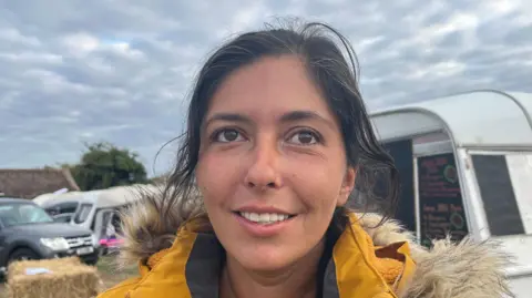 Darren Rozier/BBC A close-up image of Ellie Deacon. She is standing in front a caravan and parked cars. She has dark hair and is wearing a yellow coat.