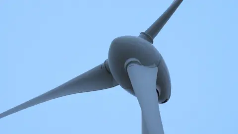 BBC A close up of the centre of a wind turbine. Three white blades are attached to the dome shaped centre which stands on top of a large mast.
