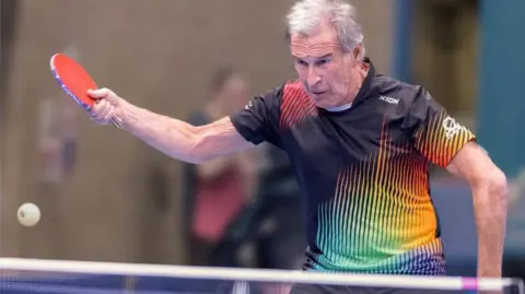 Michael Loveder A table tennis player mid-swing in a multi-coloured top, his face is a picture of concentration and the ball is on its way across the table.
