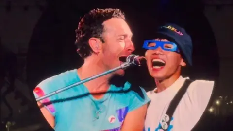 Hull KR A circular screen at a concert shows Chris Martin who has short curly brown hair and a blue top singing into a mic and looking at a man to his side. The man next to him has his mouth wide open singing and is wearing rectangular blue glasses and a Coldplay bucket hat.