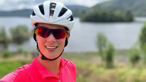 Bex Hind Bex Hind, who is wearing a white cycling helmet, sunglasses and a bright pink top, smiles at the camera. There is a lake in the background and green mountains. 