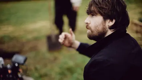 Etienne Laine A brown haired man with a beard, wearing a dark coat in a field. He looks towards a camera operator who is slightly out of shot, giving them a thumbs up. An actor holds a shovel in the background.