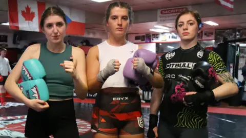 Three young women pictured in a gym. They are wearing workout clothes and are holding boxing gloves. Two have their hands raised in fists and all three have their hair tied back in ponytails.